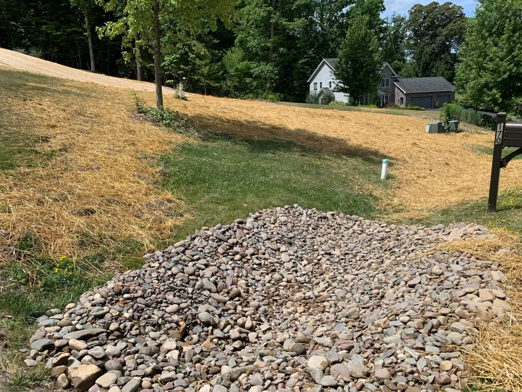 River rock apron spread across the foot of a freshly seeded and strawed slope, at the roadside end of the yard