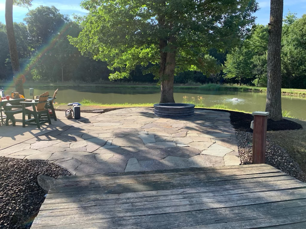 Irregular flagstone patio with a round stone fire pit and seating, laid out beside a pond under mature trees
