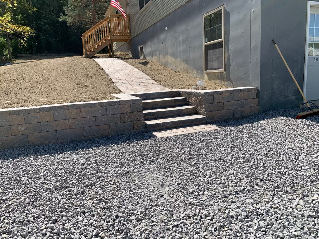 Block retaining wall with built-in steps and a paver walkway climbing to the deck of a house, with a new stone drive below