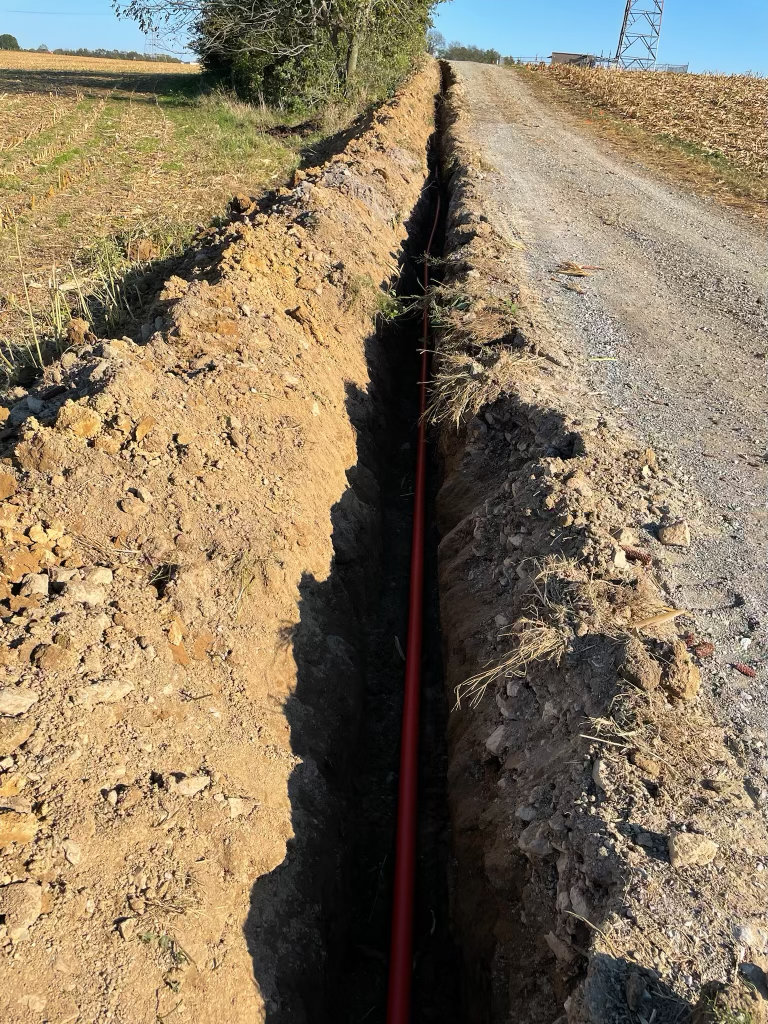 Open utility trench with conduit laid in the bottom, running alongside a gravel access lane across a field
